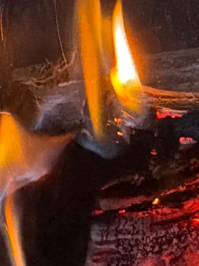 The image shows a close-up view of a fire burning in what appears to be a fireplace or bonfire. Flames in varying shades of orange and yellow are the dominant feature, with some areas of white hot intensity. Embers and glowing coals are visible within the dark, textured background, which suggests burning wood or logs. Wisps of smoke or ash are also present, contributing to the dynamic and fiery atmosphere. The scene is dimly lit, with the fire providing the primary light source. The image does not contain any identifiable landmarks or elements that would allow for specific location information within Riverhead, United States of America.
