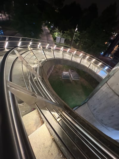 The image is an elevated, high-angle shot of a modern, illuminated spiral staircase at night. The staircase is made of concrete and metal, with polished stainless steel railings and glass panels. The lighting is a series of warm, embedded lights along the outer edge of the walkway, casting a glow on the steps and railings. Below the staircase, a circular area of grass is visible, containing two dark, pyramid-shaped structures. Surrounding the grassy area are trees and a paved path or road, illuminated by streetlights. In the background, the dark silhouettes of trees and distant lights suggest an urban environment. The scene appears to be clean and well-maintained. The architectural style and the presence of what appear to be decorative elements like the pyramid structures suggest a public space, possibly a park or a plaza. The image does not depict any people or animals. This location is identified as the Guggenheim Museum Bilbao, Spain.