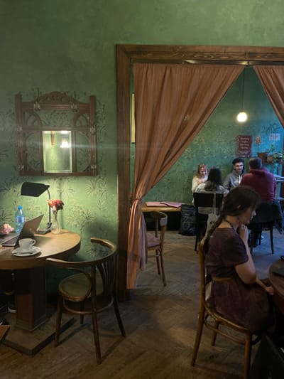 The image displays an indoor scene, likely a cozy cafe or restaurant in Kraków, Poland, characterized by a rustic and inviting ambiance. The room features a dark green wall adorned with a lighter green stenciled or painted leafy pattern, and a wooden parquet floor.
In the immediate foreground, a person's hand is visible next to an open laptop on a round wooden table. Also on this table are a clear plastic water bottle, a white ceramic cup and saucer, and a small vase holding red flowers. A black table lamp provides focused light, and a distinctive bentwood chair is positioned nearby.
A wooden partition, draped with light brown, vertically striped fabric, cuts across the middle of the frame, dividing the space. Behind this partition, a woman with braided hair is seated on a wooden chair, facing away from the camera, wearing a dark purple top or dress.
Further in the background, beyond the partition, several other people are gathered at a table. A man with dark hair and a beard, wearing a grey hoodie, is seen smiling. Beside him, a woman with light hair is also smiling. Another person, seen from the back in a reddish top, completes this group. They appear to be engaged in conversation or enjoying their time together.
The green wall in the background is further decorated with an ornate wooden mirror frame and several framed signs. One prominent sign, located near a ceiling light, displays partially legible Polish text, which includes "Puding," "Maslenka," "Syrnik," "Bifstek," and "Kartoska." These words suggest menu items such as pudding, butter dish, cheesecake, beefsteak, and potato, confirming the establishment's location in Poland. Potted plants are also arranged on a shelf on the back wall.
The lighting is artificial, suggesting the scene takes place in the evening or a dimly lit interior, contributing to a warm and intimate atmosphere. The expressions of the visible individuals convey a sense of relaxation and pleasantness.