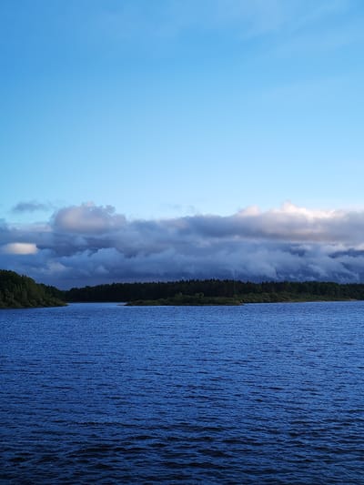 The image depicts a body of water with a dark blue hue, possibly a lake or a wide river. The water surface is rippled, suggesting a gentle breeze. The horizon line is marked by a dense forest of dark green trees. A band of low, heavy clouds hangs in the sky above the forest. The clouds are predominantly grey and white, with hints of sunlight breaking through. The upper portion of the image is a clear, light blue sky. The scene appears calm and remote. There is no indication of it being in the city of Edoma, Russia.