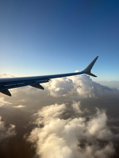 The image shows an airplane wing and tail fin extending against a backdrop of blue sky and clouds. The wing and tail fin are silver or gray in color. The sky transitions from a clear blue to a lighter hue near the horizon. The clouds vary in density, some appearing as puffy formations and others as darker, shadowy masses. The location is not evident in the image. The city, state, and country is Unknown, United States of America.