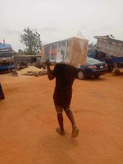The image depicts an outdoor scene in Awkuzu, Nigeria, characterized by reddish-brown earth and an overcast sky. In the foreground, an adult male, viewed from behind, walks towards the right side of the frame. He is wearing a dark t-shirt, dark shorts with light-colored cuffs, and light-colored flip-flops. He is manually carrying a large, rectangular object on his left shoulder. The object is primarily dark-colored, wrapped in clear plastic film over beige packaging, and features visible orange and white markings on its side. A label with "148" is affixed to the beige packaging, and a white, conical element protrudes from the top-right of the packaged item.
The background reveals a working yard environment. To the left, the front portion of a blue heavy-duty truck, marked with "BEST LEVEL," is visible, with a motorcycle parked in front of it. In the mid-ground, a dark blue sedan is parked, facing slightly left. To the far right, a dump truck with its bed in a raised position is partially visible. The ground is unpaved and scattered with piles of reddish-brown sand, various discarded mechanical parts, and metal debris. A large green tree stands in the distant background, flanked by several small, simple structures, likely made of corrugated metal. The scene portrays the manual transport of a packaged item within an active outdoor facility.
