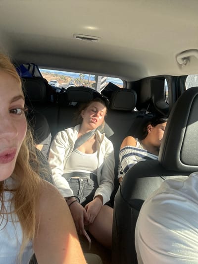 Three young women are in a car. Two are reclined asleep in the back seats. The first has dark hair and a striped shirt. The second has lighter hair, a white tank top, and a white jacket. She is wearing a seatbelt and holding a phone. The third woman, who seems to be taking a selfie, has long reddish-blonde hair and is wearing a blue shirt. The view out the window shows a sunny coastline.