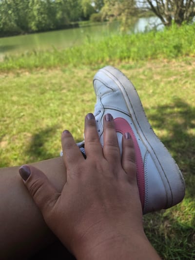 A white leather shoe with a pink Nike swoosh is resting on a person's leg, with their hand covering the shoe. The person is sitting in a grassy area with a body of water visible in the background. The location appears to be a park or natural area with trees, similar to what might be found in Epping Forest, United Kingdom.
