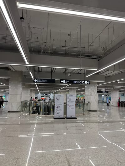 This image shows a modern, well-lit subway station in Liwan District, China. The floor is a polished, light-colored tile with reflective qualities, and lines of white light are embedded in the floor. The ceiling is concrete with exposed conduits and pipes, and large, bright fluorescent light fixtures are suspended from it.

There are several directional signs with Chinese characters and English text. One sign indicates an "Accessible Elevator" with an arrow pointing to the left. Another sign indicates "Exit B" with an arrow to the left, and "Exit C.D.E" with an arrow to the right.

In the middle ground, there are escalators leading down, with a few people using them. A glass railing with metal posts surrounds the escalator area. There are also kiosks or information booths visible. In the background, more signs and what appear to be ticketing machines or turnstiles are present.

One person is walking from the left side of the frame towards the center. The overall impression is of a clean, organized, and efficient public transportation hub.