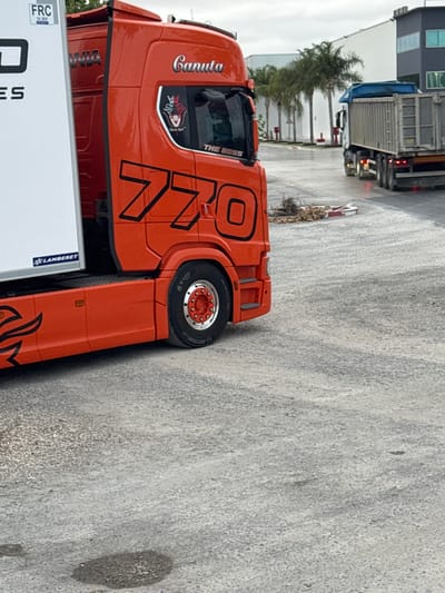 This image captures an industrial or commercial loading area in San José Obrero, Spain, under an overcast sky. The foreground is dominated by the bright orange cab of a Scania semi-truck, decorated with black graphics including the name "Canuta" and the prominent numbers "770" on its side. Below the driver's window, "THE BOSS" is written, and a window sticker displays a stylized animal with a "B" emblem. The visible rear wheel features a red hub with chrome lug nuts. A white semi-trailer, partially visible, is attached to the truck, bearing a blue "LAMBERET" logo and an "FRC 12 2023" sticker.

In the midground, a pile of natural debris, likely branches or roots, is strewn on the wet asphalt surface next to a broken red and white striped barrier. Further in the background, a white semi-truck with a grey dump-style trailer (tipper) is positioned, showing illuminated rear lights. A line of palm trees stands behind the debris, leading to a series of industrial-style buildings, including a large white structure and a darker building with reflective blue windows. The ground throughout the scene is a textured, wet asphalt or gravel surface, with a small dark puddle visible in the lower foreground.