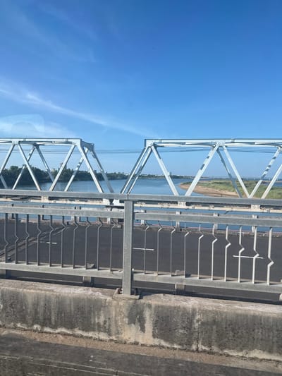 The image shows a view from a bridge, looking out over a body of water. The sky is clear and blue, suggesting it is daytime. The bridge itself is made of metal girders and has a road surface with white markings. A concrete barrier runs along the side of the road. On the other side of the bridge, there is a grassy embankment and trees lining the water's edge. In the distance, buildings can be seen. The scene appears to be a sunny day. The image is taken from a slightly elevated perspective, looking downwards at an angle. The orientation of the image is portrait, with the sky at the top and the road below. The focus is on the bridge structure and the surrounding landscape.