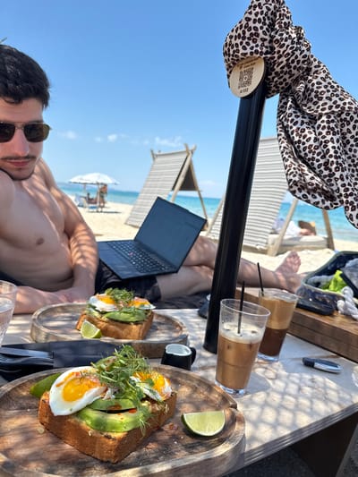 A man is relaxing on a beach in Pallini Municipal Unit, Greece, with a laptop on his lap. He is wearing sunglasses and appears to be enjoying a meal of avocado toast with fried eggs and a drink. In the background, beach chairs and umbrellas are visible, along with the ocean and a clear blue sky. A patterned scarf is tied to a black pole in the foreground, with a QR code attached to it. The table also holds two glasses of iced coffee and a small white cup.