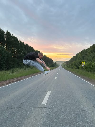A male, appearing to be in his 20s or 30s, is captured mid-air in a dynamic jump on a rural asphalt road. He is wearing a black t-shirt, light-wash denim jeans, and white sneakers. His body is arched backwards, with his legs bent and arms extended forward, creating a sense of flight. The road stretches into the distance, flanked by lush green trees and rolling hills on either side. The sky above is painted with the soft hues of a sunset or sunrise, with warm orange and yellow tones blending into pale blue and grey clouds. A speed limit sign of 70 is visible on the right side of the road, alongside a pedestrian crossing sign. The location is identified as Novaya Kazmasca, Russia, suggesting a somewhat remote, natural setting. The image's composition, with the man leaping across the road towards the horizon, emphasizes freedom and movement.