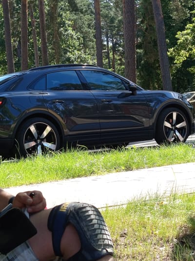 A dark gray Porsche Macan SUV is parked on the side of a paved road, with a grassy verge in the foreground. The car is seen from the side, with its rear wheel and half of the passenger side visible. The wheels have a distinctive silver and black multi-spoke design. Behind the car, tall pine trees with green foliage are lined up, suggesting a wooded area. In the lower part of the image, a person is partially visible, sitting on the grass. Their legs are covered by what appears to be protective gear, possibly for skateboarding or cycling, with a textured knee pad. The person is holding a black object, possibly a phone or a pen, and wearing a plaid shirt. The scene appears to be a sunny day, indicated by the bright lighting and shadows. The location is identified as Jūrmala, Latvia.
