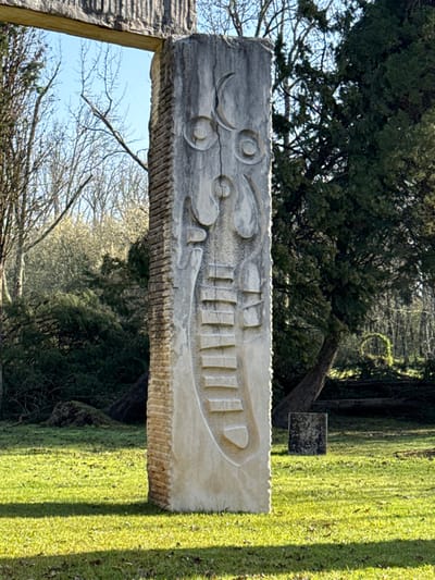 The image depicts a tall, vertical stone sculpture centrally positioned on a vibrant green grassy expanse in Mealhada, Portugal. The sculpture, primarily light grey and weathered, exhibits intricate carvings on its visible face. These carvings include a crescent shape and two circular forms with central dots near its upper section. Below, a series of abstract organic shapes transitions into approximately nine horizontal, step-like protrusions that gradually widen towards the base. A prominent vertical crack extends down the carved surface. The left side of the sculpture features a textured surface resembling stacked, rough-hewn blocks. The ground is uniformly covered in short, bright green grass, with shadows cast across its surface. In the background, diverse arboreal vegetation is present: bare-branched deciduous trees populate the left side, while dense, dark green evergreen trees occupy the right, one displaying a significantly curved trunk. A small, dark, rectangular object lies on the grass to the right of the main sculpture. The sky above is clear and light blue. No human figures are visible.