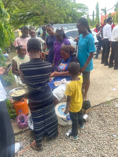 In the city of Gwagwalada, Nigeria, a group of people are gathered outdoors, possibly for a community event or a marketplace. The scene is set on a paved area with gravel surrounding it, under the shade of trees. In the foreground, a woman in a dark patterned dress is standing over a gas cylinder and a frying pan filled with what appears to be food being fried. Beside her, a young boy in a yellow t-shirt and dark pants is observing the cooking. Several other individuals, mostly women and girls, are clustered around, watching the process or engaged in conversation. Some are dressed in casual clothing, while others wear more formal attire, including white shirts and dark pants, suggesting a mix of participants. A vehicle, possibly a van, is visible in the background, along with other people walking or standing. The overall atmosphere seems to be one of social interaction and food preparation.