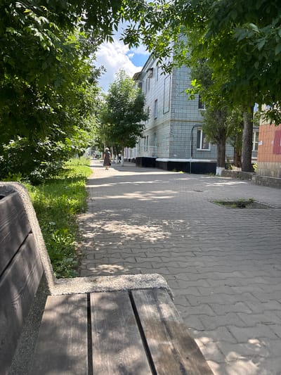 The image depicts a sun-dappled paved walkway in Gukovo, Russia, bordering a grassy area and a residential building. In the foreground, a portion of a wooden and stone bench is visible. A woman in a dark dress is walking away from the camera on the walkway. The building features light blue tiled exterior walls. Trees with green foliage line the walkway, casting shadows on the ground. The sky above is partly cloudy.