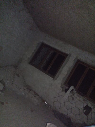 The image depicts an exterior view of a dimly lit, unfinished concrete building, likely located in Veremia, Ukraine. Two rectangular windows are visible, set into the textured concrete wall. The window frames are made of dark wood, and the panes are dark, suggesting they are either covered or unlit. The window on the left has a diamond-patterned metal grate. The ground in front of the building is rough and uneven, covered in debris. A white sheet of fabric is draped over some sort of wire mesh or fencing material in the foreground. The overall impression is one of abandonment or ongoing construction in a somber, low-light environment.