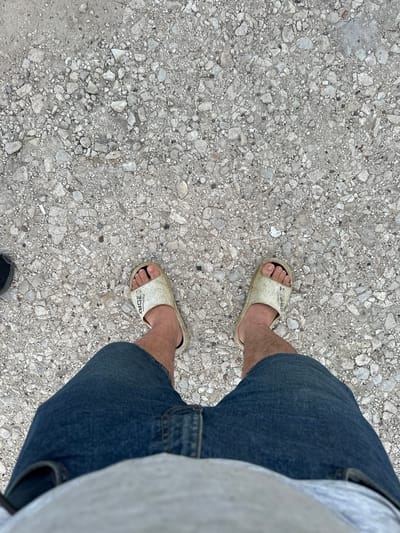 The image shows a person's legs from the knees down, wearing denim shorts and a light grey t-shirt, standing on a rough, gravelly surface. The person is wearing a pair of white slide sandals. The ground is composed of small, light-colored stones and pebbles, with some larger grey stones interspersed. The lighting suggests it is daytime, with no strong shadows visible, indicating either overcast conditions or a time of day when the sun is not directly overhead. The scene is a ground-level perspective, looking down at the person's feet and legs. There is no discernible emotional expression, but the casual attire and footwear suggest a relaxed and informal setting. Some faint text is visible on the straps of the sandals, appearing to read "FASHION 2083". The location context provided is Pîrlița, Moldova.
