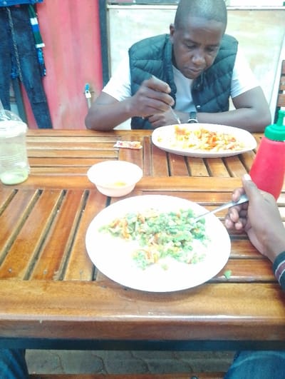 A man sits at a wooden table in a restaurant in Rungiri, Kenya. He is wearing a white t-shirt and a black puffy vest. He is holding a fork in his right hand and is eating pasta with tomato sauce from a white plate in front of him. An empty plate with remnants of pasta and tomato sauce sits on the table to his right. A small white bowl sits in front of him on the table, and a green plastic bottle filled with a red liquid sits to the right of him on the table. A second person's hand is visible in the bottom right corner of the photo, holding a fork. The table is made of wood planks with slatted sides.  An out-of-focus background shows a red wall and a blue wall covered in clothes. The location is likely a street-side restaurant with basic furnishings. The man appears to be enjoying his meal.  The photo is taken from an angle slightly below the table.  The lighting appears bright and natural.  The image is in focus throughout.  The details suggest a typical meal in a casual setting in Rungiri.  The image provides no further information regarding the identity of the individuals.  The coordinates provided (latitude: -1.25, longitude: 36.684) are located in the town of Rungiri, Kenya.