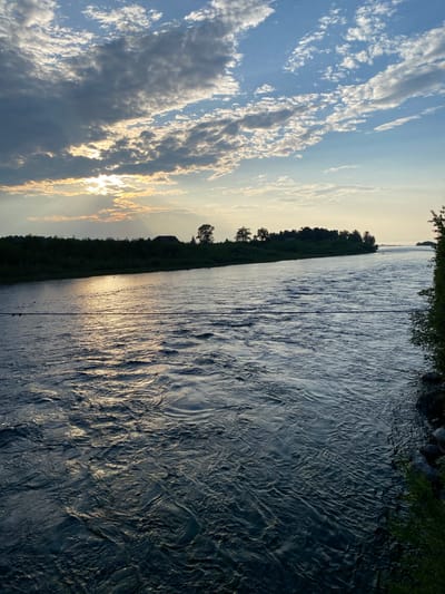 The image depicts a wide river flowing through a landscape under a partly cloudy sky. The sun is setting or rising, with rays of light piercing through the clouds and reflecting on the water's surface, creating a shimmering effect. The riverbanks are lined with dense green vegetation, and a few trees and a building are visible on the distant bank. The water itself appears to be choppy, with visible ripples and waves. There is a dark, thin line stretching horizontally across the middle of the image, possibly a wire or cable. The overall scene is serene and natural, with no people or animals visible. The location is identified as being in the city of Novosnezhnaya, Russia.