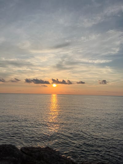 The image depicts a serene sunset over the ocean, with the sun low on the horizon casting a shimmering reflection on the water. The sky is a gradient of soft oranges, pinks, and grays, with scattered clouds. In the foreground, dark rocks are visible, suggesting a coastal viewpoint. The scene is devoid of any people or obvious man-made structures, emphasizing the natural beauty of the sunset. It is not possible to determine the specific location within Palermo, Italy from this image alone.
