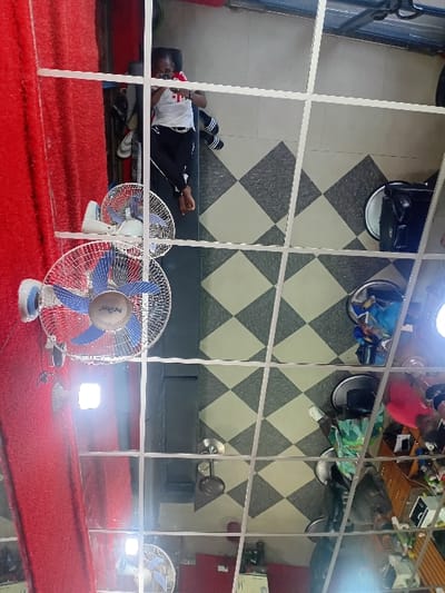 This image depicts an interior scene, reflected in a grid-paneled mirror ceiling, within a commercial establishment in Dorayi, Nigeria. The room features a floor covered with black and light-colored diamond-patterned tiles. A wall on the left side (as seen in the reflection, so the right side of the actual room) is clad in a textured red material.
Several individuals are present. In the upper left quadrant of the reflection, a light-skinned individual with short dark hair is observed lying on their back on a dark mat or bench. This person is wearing a white t-shirt with a red emblem and black athletic pants with white stripes, and is barefoot. They are holding a white smartphone horizontally, appearing to interact with its screen. Nearby, a pair of light-colored sandals and other footwear are placed.
To the right of the first individual, two white ceiling fans with blue blades are mounted, casting reflections on the mirror. Multiple bright light sources are also reflected.
In the lower right quadrant of the reflection, another individual is partially visible, wearing a green patterned top over a dark garment and white shoes, seated in a salon-style chair with a round base. Adjacent to this area, a wooden cabinet with multiple drawers holds various bottles and items, consistent with a salon or barbershop. Other salon equipment, including additional styling chairs with black seats and metallic bases, and miscellaneous objects, are distributed across the tiled floor. Another individual wearing a pink top is partially visible in the middle right, also reflected near salon equipment.