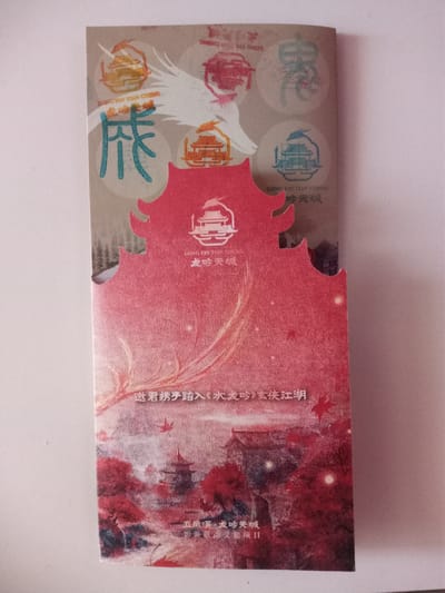 The image displays a vertical, unfolded brochure with an artistic design. The top section features circular emblems with dragon imagery and Chinese characters. Below this, a prominent red, jagged silhouette of a building is depicted, resembling a traditional Chinese temple or palace. The lower portion of the brochure shows a vibrant, painterly landscape with red trees, traditional architecture, and subtle highlights. Chinese text is present throughout the brochure. There are no people or animals visible in the image. The brochure is lying flat on a white surface. There is no indication of this image being in the city of Usman, Russia.