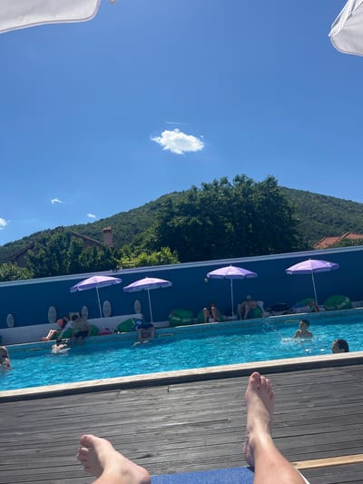 The image depicts a bright, sunny day at an outdoor swimming pool. In the foreground, a person's bare feet are visible, resting on a wooden deck. The pool itself is a vibrant blue and populated with several individuals. Some people are lounging by the poolside on beanbag chairs under purple umbrellas, while others are actively swimming or playing in the water. The background features a lush, green hillside with some trees and a glimpse of a building with a red-tiled roof and a chimney. The sky is a clear, brilliant blue with a few scattered white clouds and a faint contrail from an airplane. The setting appears to be a leisure area, likely part of a resort or hotel in the vicinity of Varvara, Bulgaria, given the context provided. The overall atmosphere is one of relaxation and recreation on a summer day.