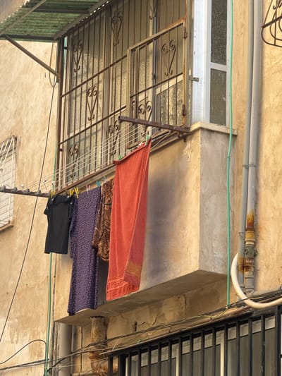 The scene depicts a view upwards towards an overhanging structure. The structure appears to be a balcony or awning with a metal frame and a translucent corrugated roof. Clothes are hanging on a line attached to the frame. The clothes consist of an orange towel, a patterned piece of fabric in brown and gold, a floral-patterned blue cloth, and a black garment. Above the structure is a concrete wall with exposed pipes and electrical cables. There is also a window with a white frame. The scene appears to be located in an urban area. While the location is stated to be Givatayim, Israel, the image does not contain any identifying elements to confirm this.