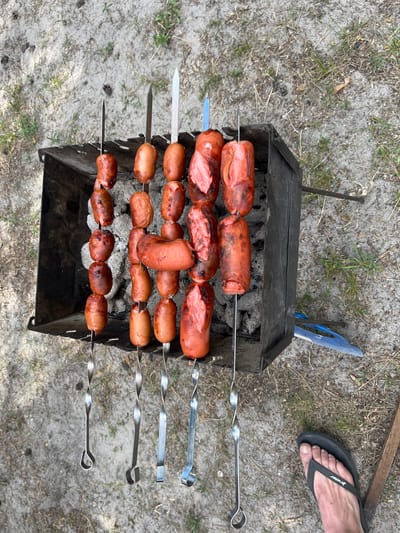 In Vyshhorod, Ukraine, an outdoor scene captures a close-up, overhead view of a barbecue. A black metal brazier, filled with coals, holds several skewers of cooked sausages and possibly tomatoes. The sausages are varied in size and color, some appear slightly charred. They are arranged vertically on metal skewers, with some hanging down. The ground around the brazier is sandy and appears to be covered with dry grass and twigs, indicating an outdoor, possibly park or backyard setting. A person's bare foot wearing a black flip-flop is visible in the bottom right corner, suggesting human presence. A blue spatula-like object is partially visible near the brazier. The overall impression is that of food preparation in an informal outdoor setting.
