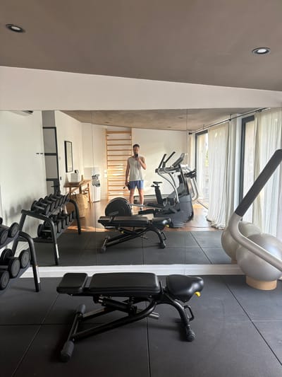 A man takes a mirror selfie in a modern home gym, reflecting a clean and well-equipped space. The gym features a variety of exercise equipment, including a weight bench in the foreground, a dumbbell rack to the left, and various cardio machines such as an elliptical and a stationary bike in the background. A large mirror covers most of the wall, reflecting the entire gym and the man. The environment appears to be a residential setting, possibly a home in Montuïri, Spain, given the natural light streaming through the sheer curtains in the background, suggesting a bright, sunny day. The man is wearing a white tank top and blue shorts, holding a phone to take the picture and a black water bottle in his other hand. The overall atmosphere is one of a private, dedicated space for fitness and wellness.