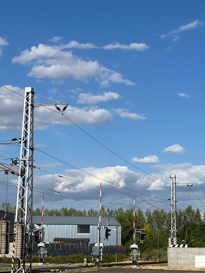 The image depicts a railway crossing with a clear blue sky filled with white, scattered clouds. In the foreground, two tall, white lattice metal poles support overhead electrical wires for the railway. These poles have various insulators and equipment attached. To the left of the image, a counterweight system is visible on one of the poles.
In the middle ground, there are multiple railway crossing gates with alternating red and white stripes. These gates are lowered, indicating the crossing is active or about to become active. Behind the gates, a corrugated metal building is visible, with stacks of black pipes or tubes on pallets in front of it. A dark fence runs along the perimeter of the building. To the right, another similar metal pole is present, supporting overhead wires. A yellow "X" shaped sign is visible on a pole, indicating a level crossing.
The environment is an outdoor setting, likely near a railway line in Újezd u Brna, Czechia. There are no people visible in the image. The overall scene is one of infrastructure related to a railway system, under a bright, partly cloudy sky.
