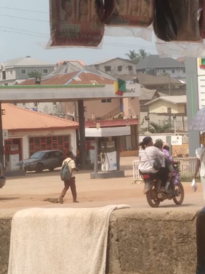 The scene depicts an active urban environment in Agege, Nigeria. In the immediate foreground, a light-colored, possibly off-white, fabric is draped over a low concrete wall, partially obscuring the bottom of the frame. Above, several retail items, likely hair products, are packaged in transparent plastic bags and suspended from an unseen overhead structure, slightly visible against the sky.

The midground features a dirt road with visible ruts and a depression. A male individual, wearing a light-colored polo shirt, dark trousers, and a blue and white patterned backpack, walks from right to left across this road. Further to the right, a motorcycle (okada) travels in the same direction, carrying three individuals: a driver, a primary passenger with dark hair, and a smaller, child-like passenger wearing patterned clothing seated behind the first. A dark-colored sedan is stationary on the left side of the road, adjacent to a building with an orange-brown roof.

In the background, a NNPC (Nigerian National Petroleum Corporation) gas station is prominent, featuring a white and green canopy structure with the corporation's logo visible. Several white and blue fuel pumps are positioned under the canopy. To the left of the gas station, a single-story building with a red roof and red-framed windows/doors is visible. Behind the gas station, a light-pink multi-story building with brown trim and exterior steps can be seen. The distant background consists of densely packed residential buildings of varying heights and colors, predominantly grey and white, with assorted roof types including corrugated metal and some flat roofs. Palm trees are interspersed among the structures, under a light blue, hazy sky.