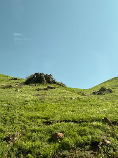 The image depicts a rural, hilly landscape with vibrant green grass covering the slopes. Several large rocks are scattered across the terrain, with one prominent cluster of dark rocks near the crest of a hill in the center. The sky is a clear, bright blue with a faint, rectangular reflection, possibly from a window, visible in the upper left quadrant. The scene appears to be a natural environment, likely outdoors, given the open sky and grassy hills. The specific location in the city of Tsovagyugh, Armenia, indicates this is a view of the Armenian countryside.
