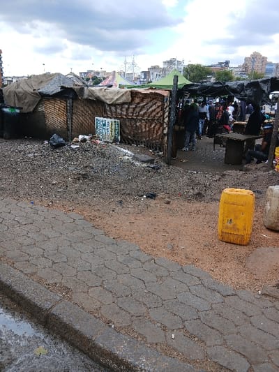 A street scene shows makeshift shacks constructed from wood and covered with tarps, positioned along a rough, dirty ground. A yellow container rests on the ground, next to an area paved with grey bricks. Further back, market stalls are visible under a black awning. The city skyline is visible in the background, partially obscured by a cloudy sky.