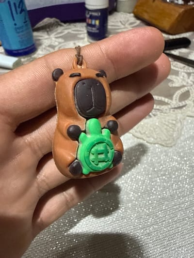 A close-up shot captures a person's hand holding a small, brown capybara figurine with a green turtle pressed against its chest. The figurine, likely made of plastic or rubber, has a simple design with a dark nose and eyes. The capybara's paws are rounded, and the turtle features a carved pattern on its shell. A thin, brown string is attached to the top of the figurine's head. The background consists of a textured, white surface with a subtle, sparkly sheen. To the left of the hand, the edge of a blue bottle with white lettering is visible, along with a blurry blue object behind it. Further back, a light brown, wooden object rests on the textured surface. The image appears to have been taken indoors, with no specific geographical indicators to suggest it is located in Hadera, Israel.
