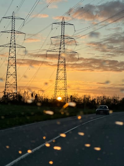 This image captures an outdoor scene at either sunrise or sunset in Marcoussis, France, characterized by a prominent sun low on the horizon. The sky displays a gradient from deep orange-gold near the horizon to a lighter blue-grey overhead, with scattered stratocumulus clouds.

Two large, lattice-steel electricity transmission pylons are centrally positioned, silhouetted against the bright sky. Multiple high-voltage power lines extend horizontally across the frame, connecting the pylons. Several spherical aircraft warning markers are visible on the lower power lines. The sun is partially visible at the horizon, situated directly beneath the rightmost pylon, casting a strong golden illumination.

In the mid-ground, a two-lane asphalt road with a broken white center line curves from the lower-center towards the right. A dark-colored sedan vehicle is observed driving away from the viewer on the right lane of this road.

A strip of dense, bare trees and shrubs forms a dark, silhouetted barrier between the road and the distant horizon, suggesting a cooler season. Green ground cover is visible to the left of the road. The immediate foreground is obscured by numerous illuminated water droplets or dew, likely on a glass surface, creating distinct bokeh effects. No human figures are present.