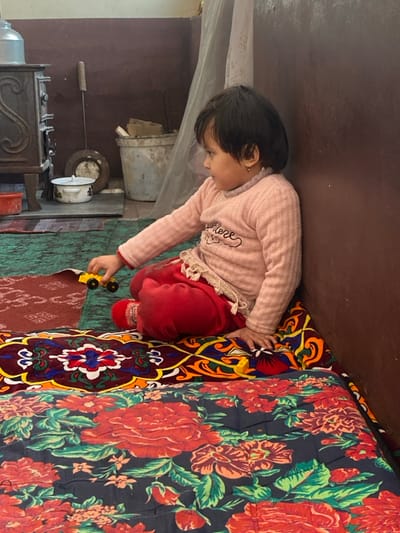 A young girl sits on a colorful floral-patterned mattress, playing with a yellow toy car. She wears a pink sweater and red pants. Behind her, an old stove sits in what appears to be a kitchen or living space. The room has a rustic feel, with a bucket and other items stored nearby. The scene captures a moment of childhood play in a simple, lived-in setting.