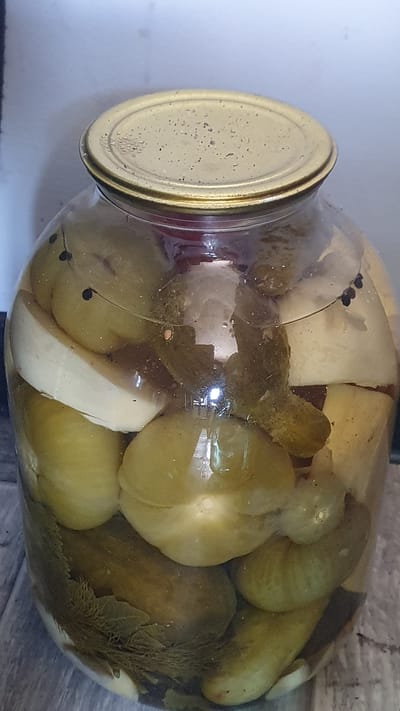 The image shows a large glass jar filled with pickled green tomatoes and cucumbers, along with what appear to be slices of garlic and dill. The jar is sealed with a tarnished gold-colored metal lid. The contents are submerged in brine. The jar is placed on a wooden surface, likely a table or shelf, with a plain, light-colored background behind it. There are no people or animals visible in the image. The scene appears to be a domestic setting, possibly a kitchen or pantry, in the city of Ust-Labinsk, Russia.