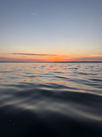 This is a vertical, eye-level shot of a tranquil body of water at sunset. The foreground is dominated by dark, rippling water reflecting the muted colors of the sky. As the eye moves towards the horizon, the water becomes lighter, reflecting the warm orange and pink hues of the setting sun. The sky transitions from a pale blue at the top to a band of soft orange and pink near the horizon, with a few wispy clouds. A thin contrail from an airplane is visible in the upper left quadrant of the sky. The distant horizon is lined with a dark silhouette of trees, with a few indistinct structures, possibly industrial, visible. The scene is devoid of people, animals, or any obvious activity. The overall impression is one of calm and natural beauty. Without additional information, it is not possible to definitively confirm the location in Salaspils, Latvia. However, the presence of a large body of water, a sunset, and distant industrial structures are all consistent with a location near the Daugava River in or around Salaspils.