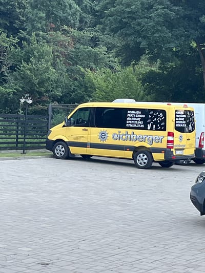 A yellow Mercedes-Benz Sprinter van is parked on a paved lot in front of a black fence and a dense green forest. The van has "eichberger" written in large, blue letters on its side, along with other text and musical notes. The license plate of the van is partially visible, reading "SV 14 SNC". There is a white van parked behind it, and the front of a dark-colored car is visible in the lower right corner of the image. The location is identified as Huși, Romania, based on a sign that reads "Formația Frații Sandu din Neamț" on the van, which is a musical group from Neamț County, Romania.