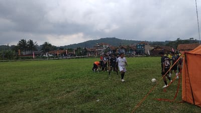 The image captures an outdoor football game or practice session on a green grassy field, set against a backdrop of a village and mountains under a cloudy sky. A group of young men, likely football players, are gathered on the field. Most players wear dark jerseys with white numbering and text, while one player is in a white jersey and another in an orange shirt is bending over. A white football is visible near a player wearing a black jersey with the number "04" on the back. Partially visible on the right is an orange pop-up tent or shelter, with red ropes extending from it, suggesting a temporary goal or sideline structure.
In the mid-ground, a village stretches out with numerous houses featuring roofs of various colors like red, blue, and brown. Palm trees and other foliage are scattered throughout the village. Colorful banners or flags (pink, orange, purple) are strung along what appears to be a fence line separating the field from the village houses. The far background is dominated by a range of forested mountains, giving the scene a rural, natural feel. The sky is heavily overcast with grey clouds, indicating a cloudy day with diffused lighting. The overall impression is a casual sporting event in a picturesque, mountainous village environment, consistent with the Panjalu, Indonesia location.