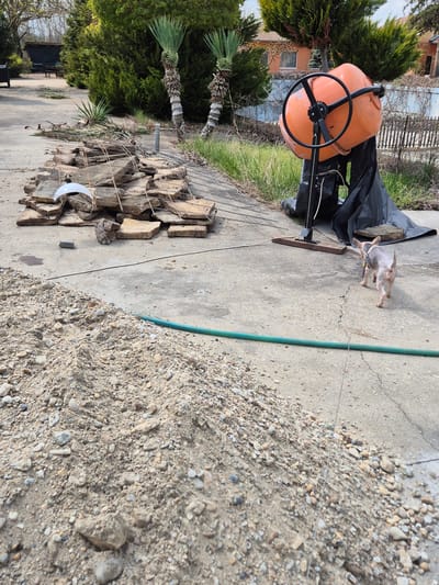 The image shows an outdoor scene in Pavlikeni, Bulgaria. The setting is a concrete-paved area. An orange concrete mixer with a black wheel sits near a pile of stacked stones and a pile of sand. A small dog stands near the mixer. A green hose runs across the concrete. There are bushes and trees in the background. A partially visible building can also be seen in the background.