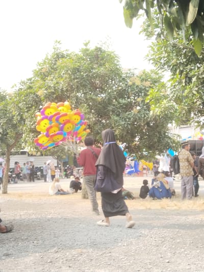 In Plosorejo, Indonesia, an outdoor scene unfolds under a bright sky, likely during the day. The ground is covered with gravel and dry grass, with several trees providing shade. In the foreground, a person wearing a black hijab and a long skirt walks away from the camera. To the left, a man in a red shirt holds a large bunch of colorful balloons shaped like flowers with smiley faces. In the midground, various individuals are scattered, some sitting on the ground, others standing. A group of people are gathered near some parked motorcycles. Further in the background, there are structures that appear to be tents or stalls, and a large black speaker system. The overall atmosphere suggests a public gathering or event.