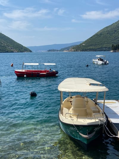 The image depicts a serene view of the Bay of Kotor in Perast, Montenegro, on a clear, sunny day. In the foreground, a green and cream-colored motorboat is docked at a wooden pier. The boat has the registration "221 KT" visible on its hull. Further out in the water, a red passenger boat with a white canopy is anchored, with a single individual seated inside. Another smaller white boat with a canopy is visible in the background, with a person swimming or diving nearby. The water is a brilliant turquoise, revealing the rocky seabed beneath its surface. Lush green mountains rise steeply on either side of the bay, framing the scene. The sky is a vibrant blue with scattered white clouds. The overall atmosphere is one of tranquility and natural beauty.