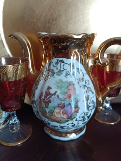 The image shows a close-up of a decorative pitcher with a painted pastoral scene, surrounded by ornate gold-rimmed glasses filled with a red liquid. The pitcher is white with a gold trim and features a painting of a man playing a guitar for a woman sitting with a basket of flowers. The glasses are made of ruby-red glass with intricate gold patterns. The objects are placed on a dark wooden surface, and a gold-colored tray or platter is visible in the background. The lighting suggests an indoor setting, possibly a display or a table setting. The scene does not provide any specific information about the location in Évreux, France.