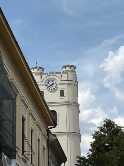 The image shows a view of the Reformed Great Church of Debrecen, Hungary, from a low angle looking upwards. The church features a prominent clock tower with a white facade and a circular clock face. The clock shows the time as approximately 9:55. In the foreground, a yellow building with ornate window frames is visible, partially obscuring the view of the church. The sky is blue with scattered white clouds, and the top of a green tree can be seen on the right side of the image. The scene appears to be taken on a bright, partly cloudy day.
