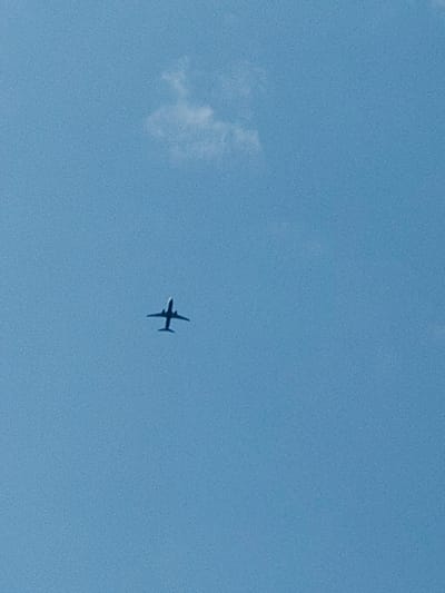 The media file shows a silhouette of a passenger airplane flying across a clear blue sky. A small, wispy white cloud is visible in the upper left portion of the image. The airplane is positioned in the lower center of the frame, with its wings outstretched and its body clearly defined against the sky. The image appears to have been taken during the daytime, with bright daylight casting a clear view of the sky. There is no visible text in the media file. The location context is Łomna-Las, Poland, which suggests the photograph was taken in that region. The scene is serene and common, depicting a typical aerial view of a plane in flight.