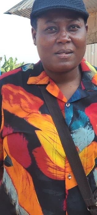 This image depicts a single dark-skinned individual, appearing to be an adult, captured in a chest-up portrait shot in an outdoor setting within the city of Alayi, Nigeria. The individual wears a black baseball cap and a collared button-up shirt featuring an abstract, multi-colored pattern on a black background, with prominent hues of red, orange, yellow, and blue. A dark strap, possibly from a bag, crosses diagonally from the individual's left shoulder towards the lower right. A small, silver-colored stud earring is visible in the individual's left earlobe. The individual's expression is neutral, with direct eye contact or a slight gaze to the right of the camera. The background is moderately blurred but reveals a light-colored sky, a section of a corrugated metal roof, and green foliage, indicative of a daytime, possibly semi-urban or residential environment. No discernible interactions or specific actions are observed beyond posing for the photograph.