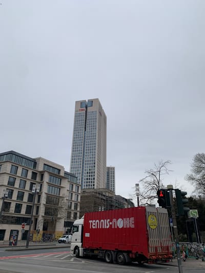 The image shows a red semi-trailer truck with the words "TENNIS-OHNE" in white letters on the side. It is stopped in the middle of an intersection in Frankfurt am Main, Germany. The truck is facing the camera and to the left, the road ahead is clear. The road behind the truck is also clear. The truck is facing a large building with "UBS" in red letters visible on the front of the building. There are two traffic lights on the right side of the intersection. The first is red, the second is green. There is also a traffic light on the left side of the intersection. There is a bird in the sky. The truck is on a city street with sidewalks on both sides. There are trees and other buildings visible in the background. Several cars are parked on the side of the road. The image is taken on a cloudy day.