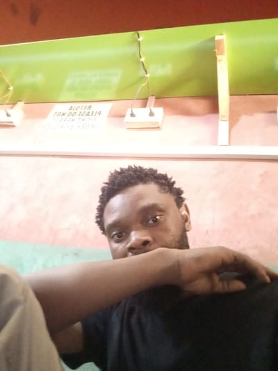 The image captures an indoor scene in Magboro, Nigeria. A Black male individual is visible from the chest up, positioned centrally. He has short, dark, textured hair and a beard, wearing a dark (likely black) t-shirt. His right forearm is raised horizontally across his upper chest, partially obscuring his lower face. His gaze is directed towards the camera. The background wall is painted with a lower section of reddish-brown/pinkish-tan and an upper horizontal stripe of lime green. Above the green stripe, a darker, possibly maroon, surface is visible. Two surface-mounted white electrical wall sockets are present on the reddish-brown wall, below the green stripe, with white wiring extending upwards. Between these sockets, a rectangular white sign displays mirrored black text. When un-mirrored, the text reads: "BETA ALERT PLEASE DO NOT [incomplete word, possibly "LOOK"] THROUGH THIS ORDER BY 09094". A white horizontal trim piece runs along the lower portion of the visible wall, and a vertical wooden plank is visible on the far right.