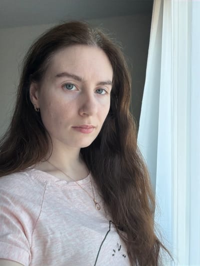 A frontal portrait shows a female subject, approximately 20-30 years old, situated indoors in Yanino-1, Russia. The subject has long, dark brown hair with visible grey strands at the roots, parted centrally. Her skin is fair, and she possesses light blue-green eyes, maintaining a direct gaze towards the viewer with a neutral expression. She is wearing a light pink short-sleeved t-shirt, featuring a partial graphic design on the lower left. Accessories include small, silver-toned hoop earrings and a delicate gold-toned chain necklace with an oval pendant. The background on the left consists of a plain dark grey wall, while the right side is dominated by a sheer white curtain, indicating a window and serving as the primary light source for the scene. No other individuals or significant objects are present within the frame.