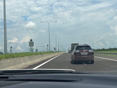 The image shows a view from inside a vehicle traveling on a multi-lane highway. The road is paved, with clear white lane markings. On the left side of the road, there is a concrete barrier with yellow reflectors. Beyond the barrier, there is a grassy verge and then more infrastructure including traffic lights and signs. Tall streetlights line the highway. In the foreground, the dashboard of the vehicle is visible. On the road, several vehicles are present, including a maroon Lexus RX 400h with license plate "CH 555-SO", a large truck, and other cars. The sky is partly cloudy with white clouds scattered across a blue sky. The environment appears to be a rural or semi-rural setting, possibly on the outskirts of a city or town. The location is identified as სუფსის თემი, Georgia.